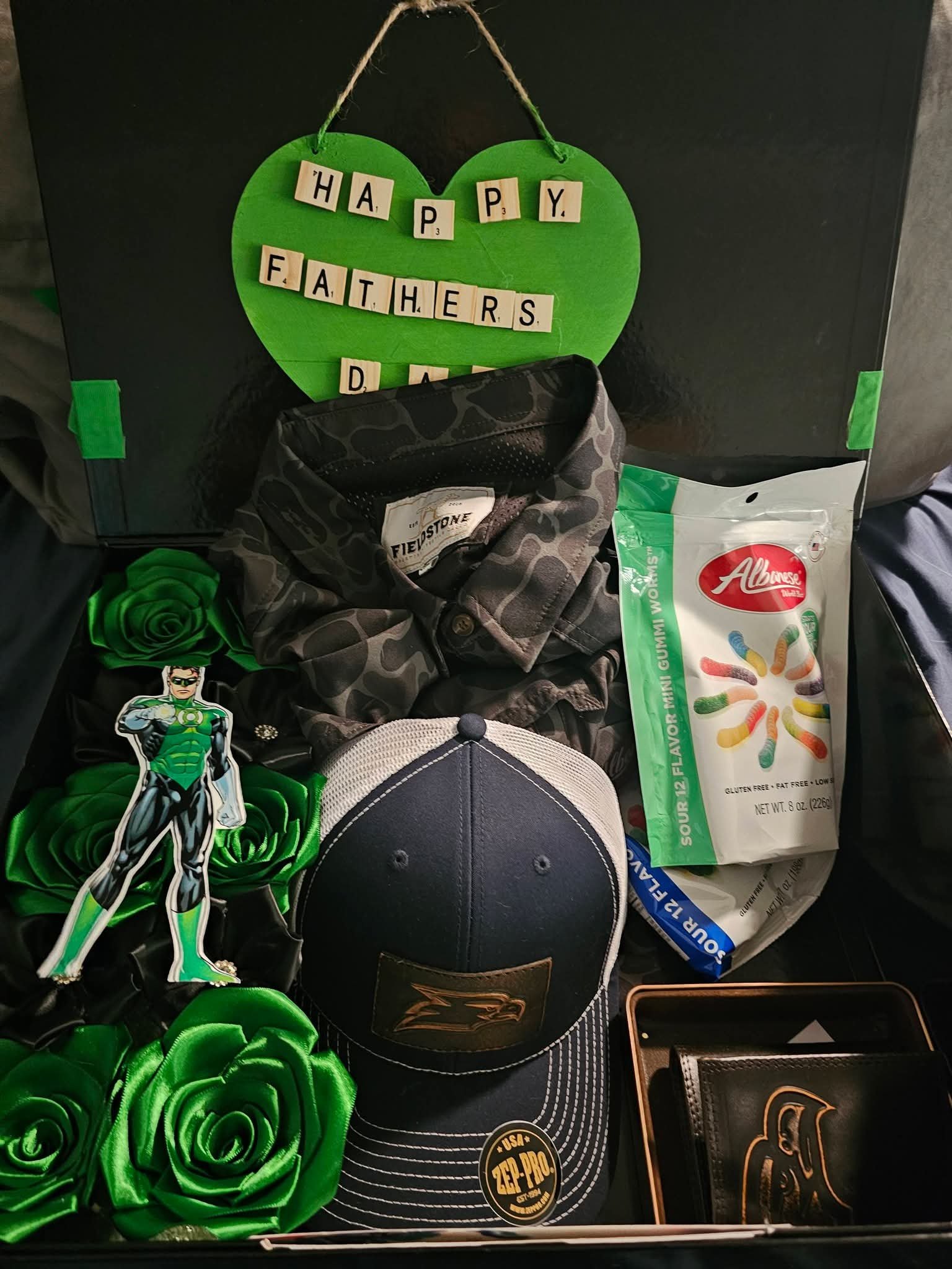 Open Happy Fathers Day gift box lined with green ribbon roses around a camo shirt, cap, wallet and candy